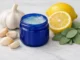 Frasco azul de ungüento mentolado junto a ajo y limón sobre mármol, ilustrando usos alternativos caseros